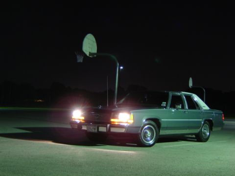 1988 Ford Crown Victoria LTD - A green sedan sits parked on a court at night, illuminated by its own headlights.