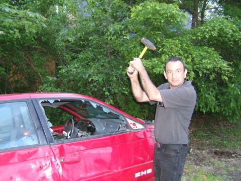 1998 Ford Taurus - A man prepares to smash a red 1998 Ford Taurus with a hammer, its windows already broken.