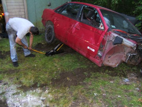 1998 Ford Taurus - A person is using a car jack to lift a damaged red sedan, which is missing its front bumper and fender.