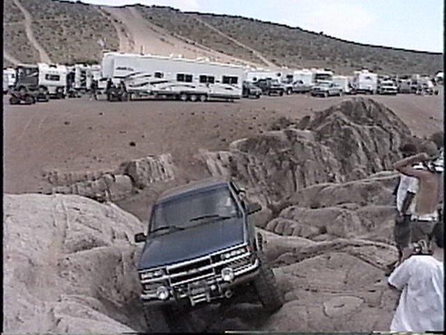 Dark blue pickup truck navigating rocky terrain. Several RVs and trailers are parked in the background