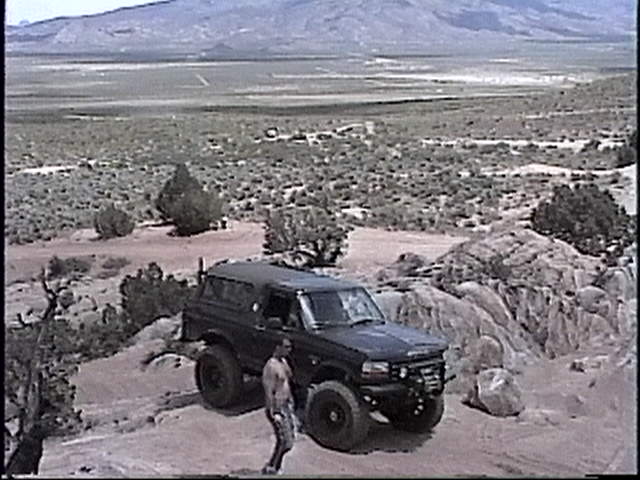 Black Ford Bronco parked on rocky terrain with a man standing nearby. Mountains in background
