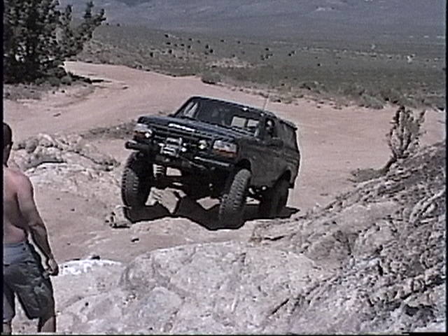 Black Ford Bronco climbing over rocks on a dirt trail. A person stands to the left