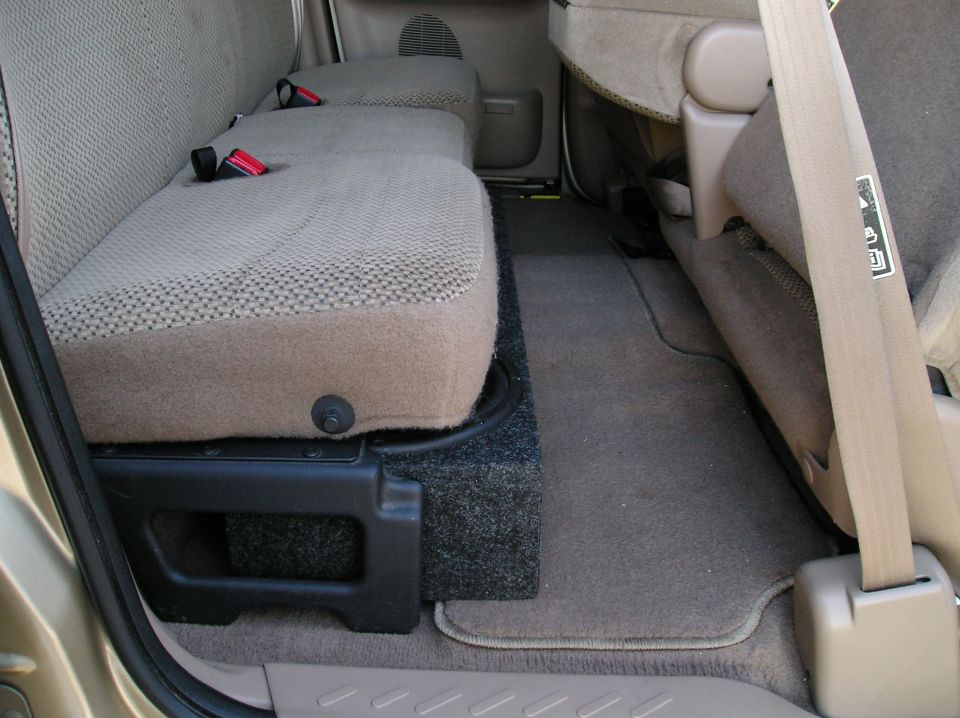 Rear cab interior of a pickup with tan cloth seats and a black carpeted subwoofer box under the rear bench