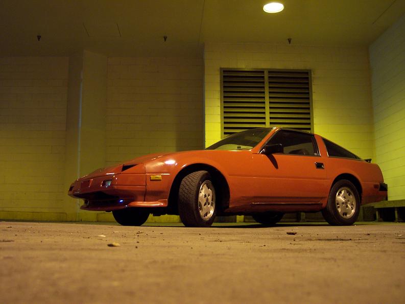Orange-red Nissan 300ZX parked at night under a light beside a louvered vent and wall