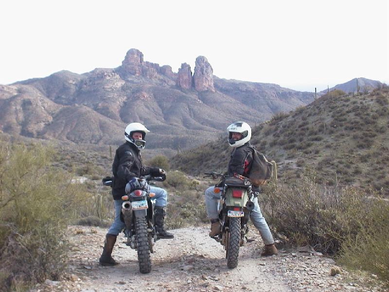 Two helmeted riders on dual-sport motorcycles on a desert trail with rocky peaks behind