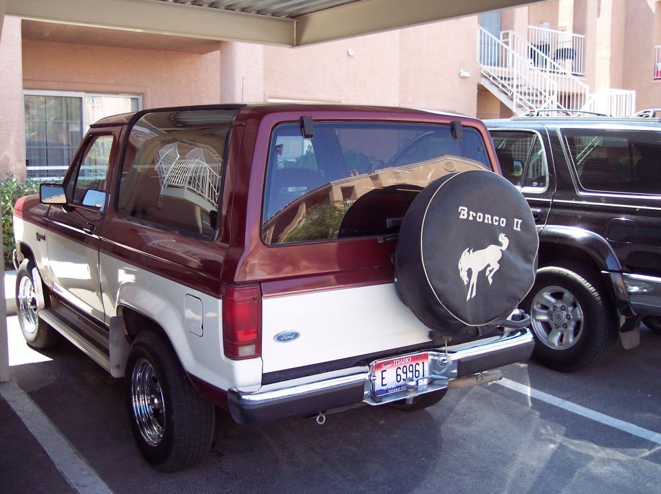 Two-tone red and white Ford Bronco II with spare tire cover, parked next to a black SUV
