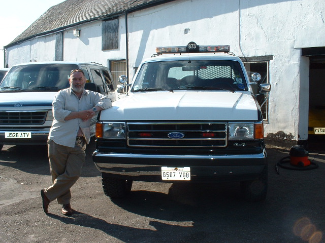 Man leaning on the front of a white Ford 4x4 with roof light bar and UK plates outside a white stone building
