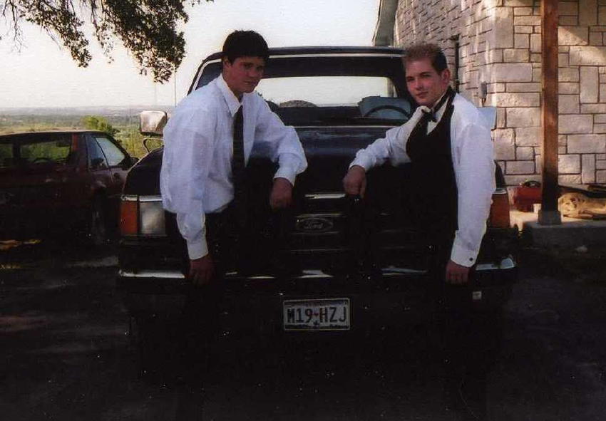 Two young men in formal attire lean on the tailgate of a dark Ford, plate M19-HZJ