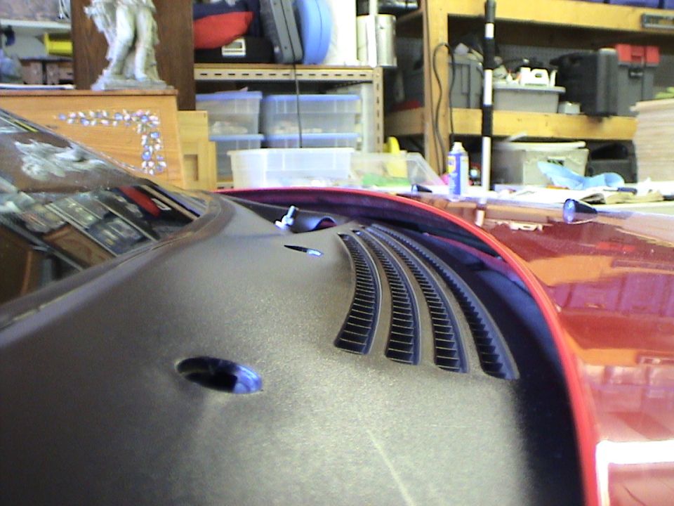 Car hood vent with horizontal slats and a dark gray textured finish. A red car is visible to the right