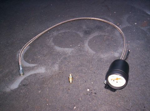 1989 Ford Bronco II - Gauge with braided hose and fitting on asphalt, likely for tire pressure or fluid monitoring.