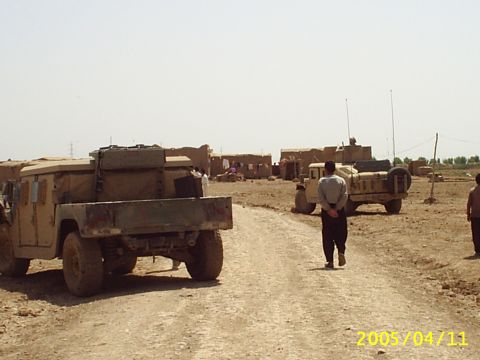 1988 Mercury Sable - Two military vehicles are visible on a dirt road in a dusty, arid environment with buildings in the background.