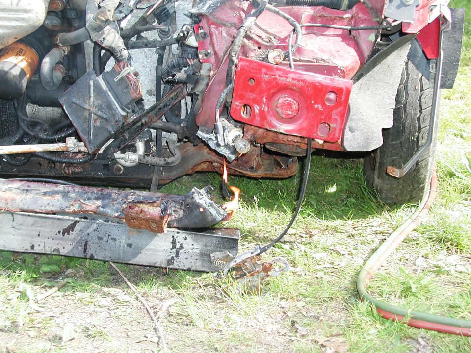 Damaged front end of a vehicle with visible engine components and a small fire burning on a metal beam