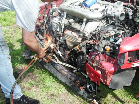 1992 Ford Taurus - A person uses a torch to cut into the engine compartment of a red car, revealing extensive damage.