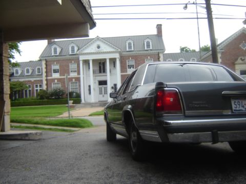 1988 Ford Crown Victoria LTD - A dark 1988 Ford Crown Victoria LTD is seen from the rear, parked in front of a grand brick mansion.