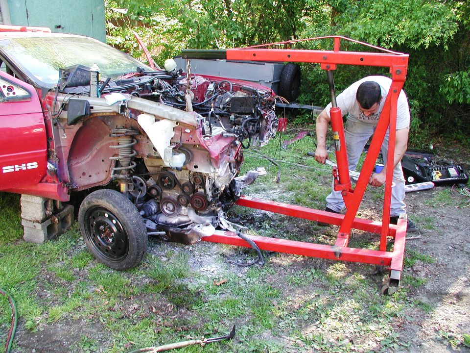 Guy using an engine hoist to pull the engine out of a red car with its front end dismantled