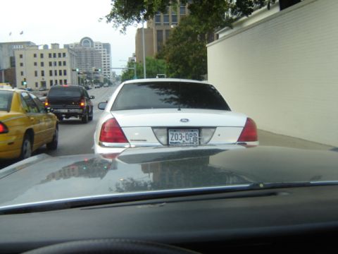 1988 Ford Crown Victoria LTD - A white sedan is seen from behind in traffic, with other vehicles on the road and buildings in the background.