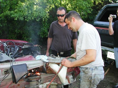 1992 Ford Taurus - Two men are working on the damaged front end of a red car, with smoke emanating from the engine compartment.