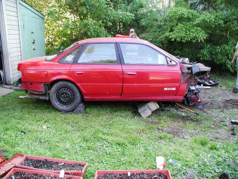 1992 Ford Taurus - A red sedan, possibly a 1992 Ford Taurus, sits in a yard with significant front-end damage and missing parts.