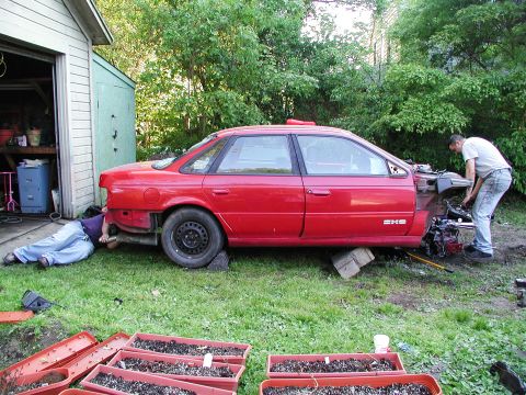 1992 Ford Taurus - Two individuals work on a red 1992 Ford Taurus sedan that has had its front end disassembled.