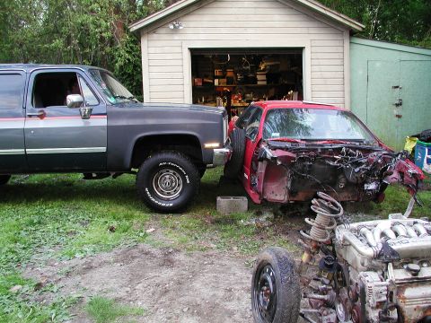 1992 Ford Taurus - A severely damaged red car sits in a yard next to a large SUV, with its engine and suspension components visible.