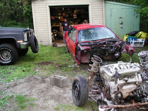 1992 Ford Taurus - A red 1992 Ford Taurus sits disassembled in a yard next to a black truck, with its engine and suspension components strewn about.