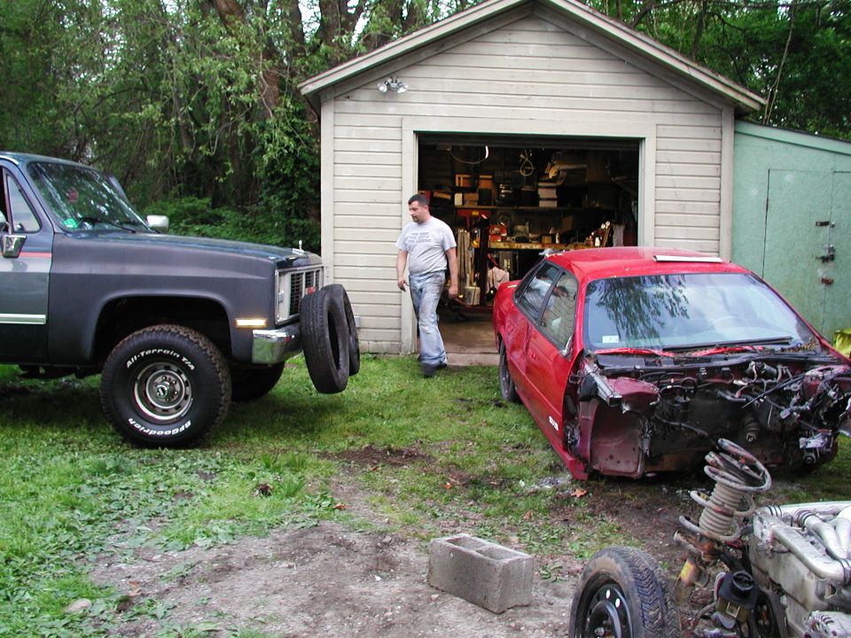 Red car stripped of its front end sits next to a dark pickup truck with two tires mounted on its bumper in a yard