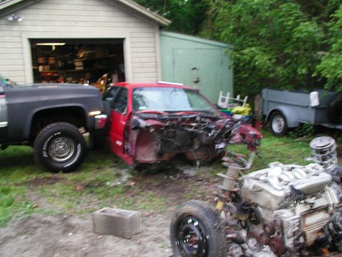 1992 Ford Taurus - A red car, stripped of its components, sits in a yard next to a pickup truck and a trailer.