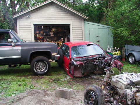 1992 Ford Taurus - A red 1992 Ford Taurus sedan with a damaged front end is being towed by a dark gray pickup truck in front of a garage.