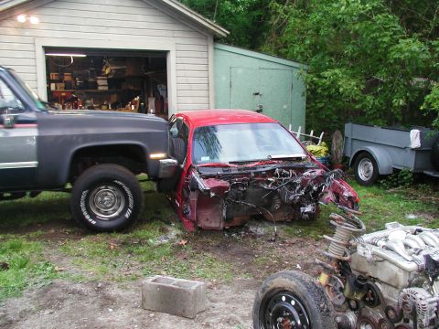 1992 Ford Taurus - The front end of a red 1992 Ford Taurus is heavily damaged and missing components, parked next to a pickup truck.