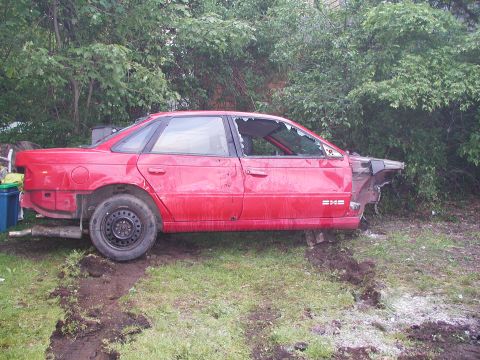 1992 Ford Taurus - Damaged 1992 Ford Taurus sedan sits in a yard with broken windows and missing front end.