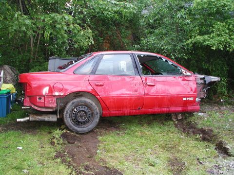 1992 Ford Taurus - A damaged red sedan sits in a grassy area with trees in the background.