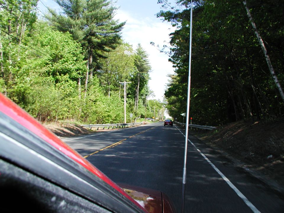 View from a car driving on a two-lane road with trees lining both sides; a single car is visible in the distance