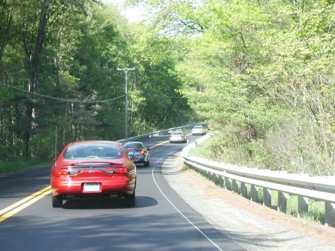 1992 Ford Taurus - A red sedan travels down a tree-lined road, followed by other vehicles.