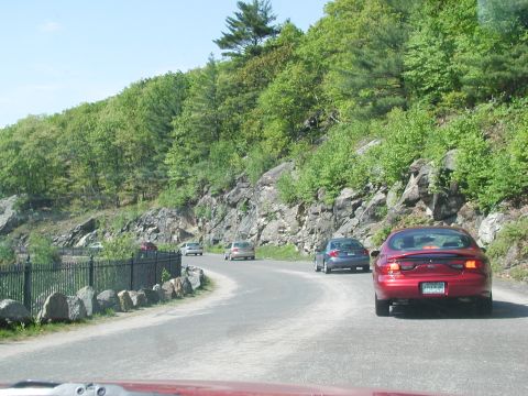 1992 Ford Taurus - A red sedan drives along a scenic, winding road bordered by trees and rocky terrain.