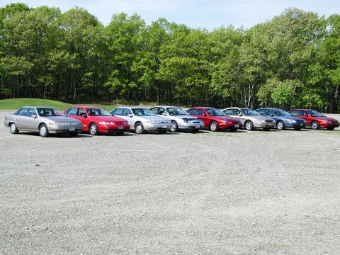 1992 Ford Taurus - A row of sedans, including a silver 1992 Ford Taurus, are parked on a gravel lot with trees in the background.