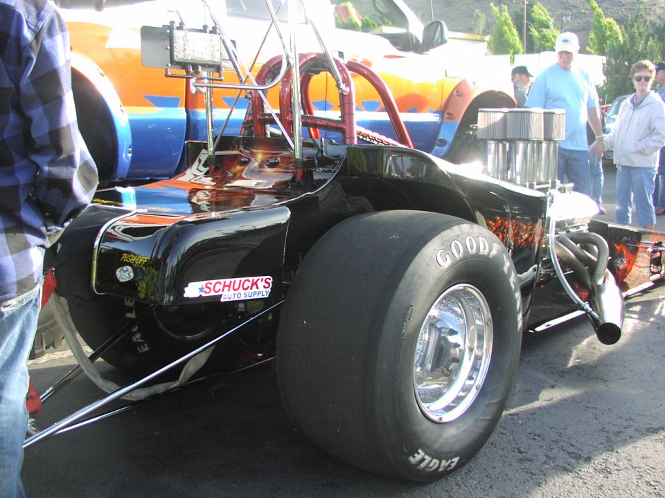 Dragster rear view with large Goodyear Eagle tire, Schuck's Auto Supply sticker, and orange and blue striped truck in background
