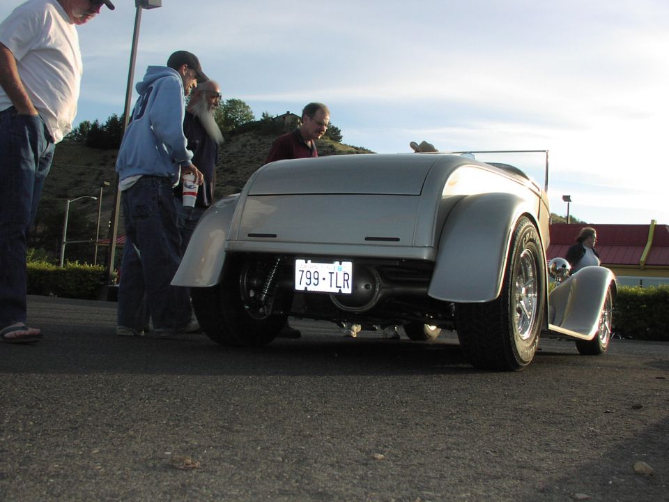 Silver hot rod with custom rear end and license plate 799-TLR. People are looking at it