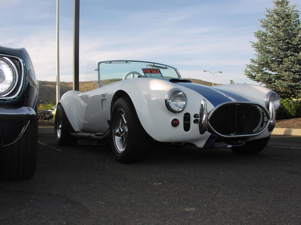 White AC Cobra replica with blue stripes, parked next to another car. Has a 'FOR SALE' sign in the window