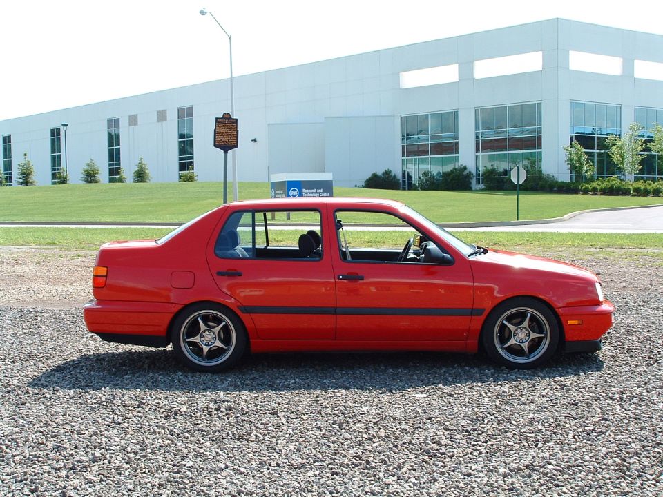Red sedan parked on gravel lot, side view. Building in background