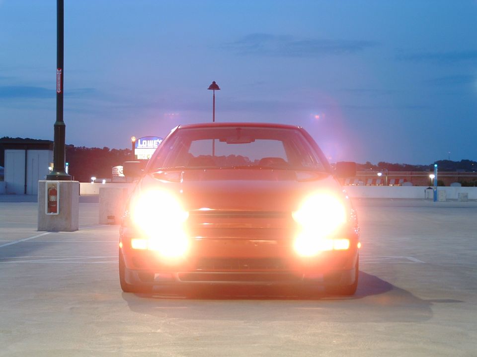Red car parked in a lot at dusk, headlights on