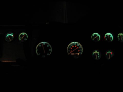 1990 Ford Bronco - vehicle's aftermarket gauge cluster glows green and red in darkness, showing a speedometer, tachometer and auxiliary dials.