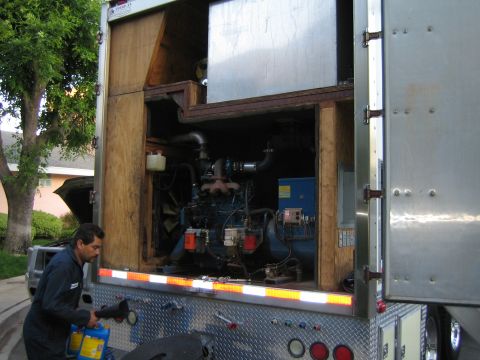 1990 Ford Bronco - man in coveralls works at the back of a trailer whose open compartment houses a diesel generator, on a tree-lined street at dusk.