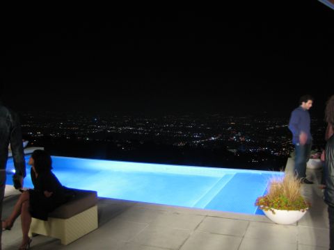 1990 Ford Bronco - blue-lit infinity pool on a hillside terrace overlooks distant city lights at night, with several people standing and seated nearby.
