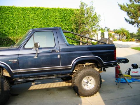 1985 Ford Bronco - A dark blue Ford Bronco, modified with a roll cage and off-road tires, is parked outdoors.