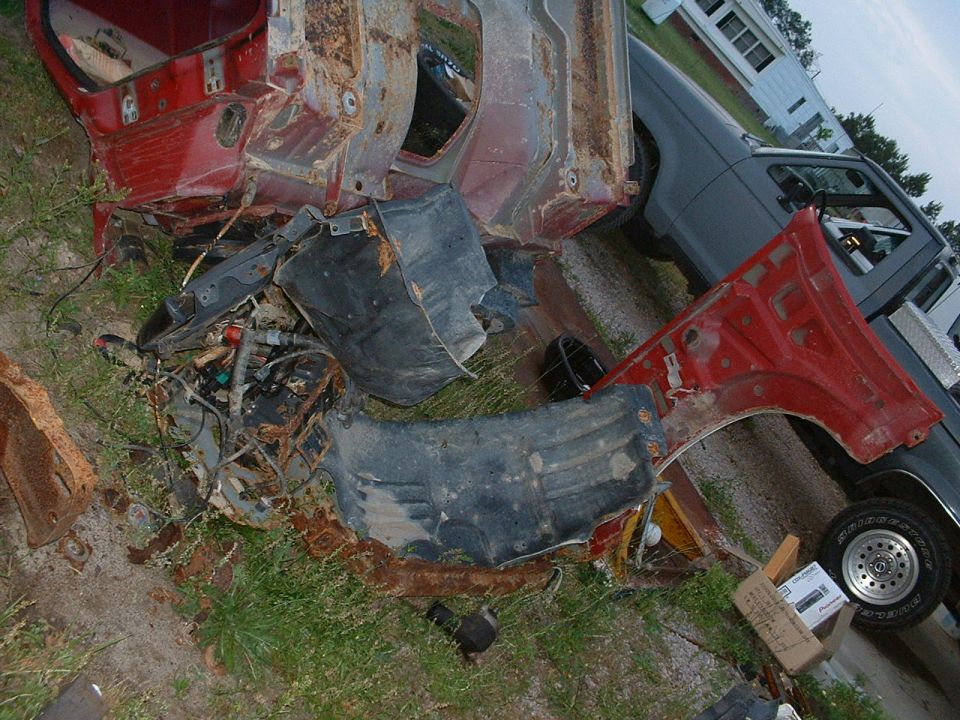 Disassembled red vehicle body and engine parts scattered on grass next to a gray pickup truck