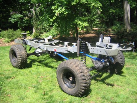 1978 Ford Bronco - A bare rolling chassis frame sits on grass in a wooded yard, showing the ladder frame, front and rear axles with coil springs, blue-painted…