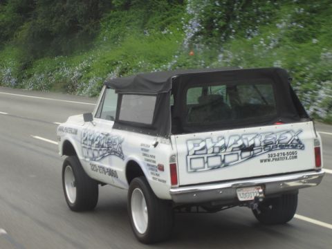 1990 Ford Bronco - lifted early-1970s SUV with a black soft top and 'PHATFX' body graphics is captured from behind moving on a highway.