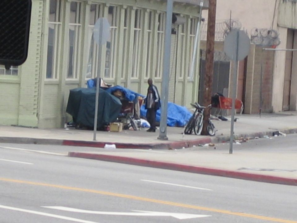 Homeless encampment with tarps and a bicycle on an urban sidewalk beside a green building