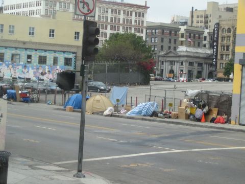 1990 Ford Bronco - Tents and belongings line a downtown sidewalk across from a fenced lot, with the historic Hotel Barclay visible behind.