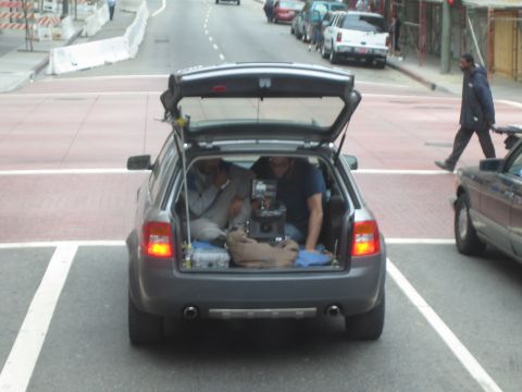1990 Ford Bronco - gray wagon parked diagonally on an urban street with its rear hatch open, loaded with camera equipment and two people inside.